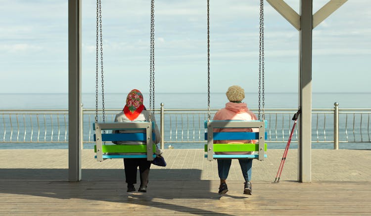 Back View Of Senior Women Sitting On Swings And Looking At Seascape