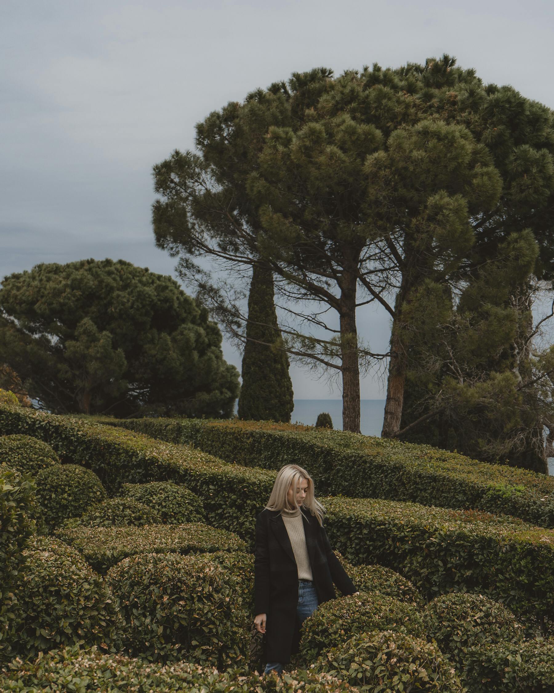 Woman in black coat walking through a lush botanical garden with tall trees.