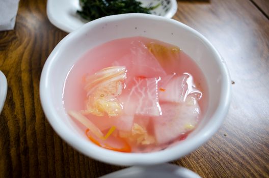A close-up of a traditional Korean pink radish pickle dish served in a white bowl on a wooden table.