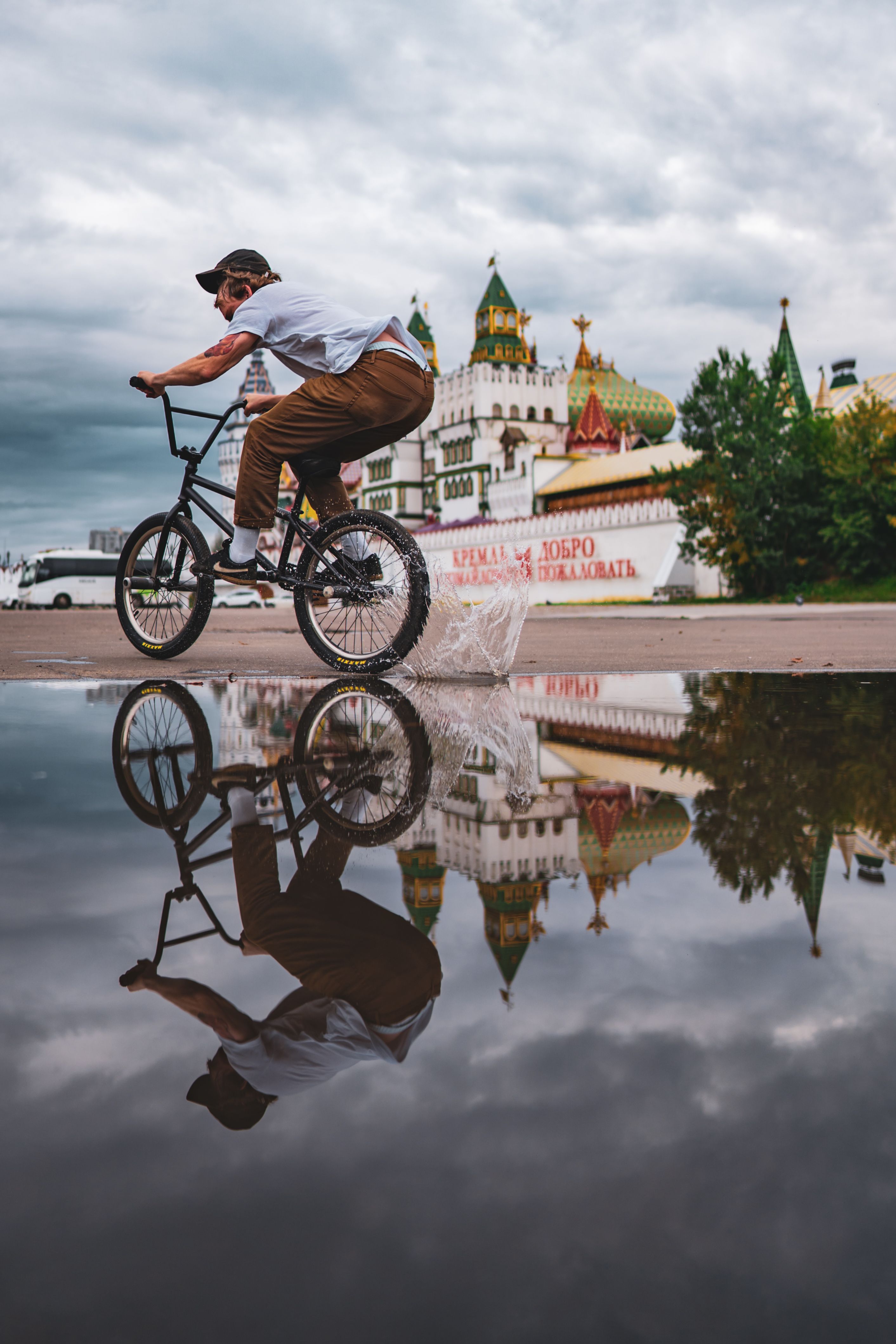 A BMX biker performs stunts near a historic Russian building, with reflections in a puddle.