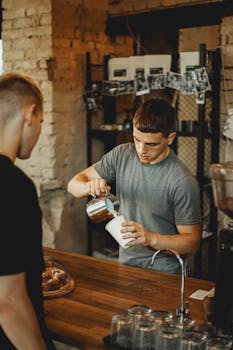 A barista pours milk into a coffee cup in a cozy, rustic cafe setting, focusing on latte art.
