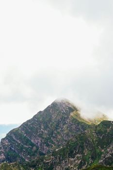 A scenic view of a rocky mountain peak partially covered by clouds against a bright sky.