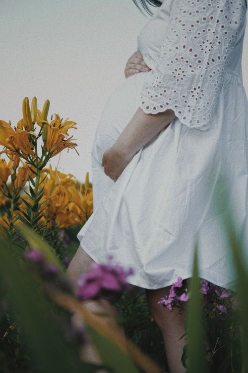 Free Side view of a pregnant woman in a white dress walking among vibrant flowers outdoors. Stock Photo