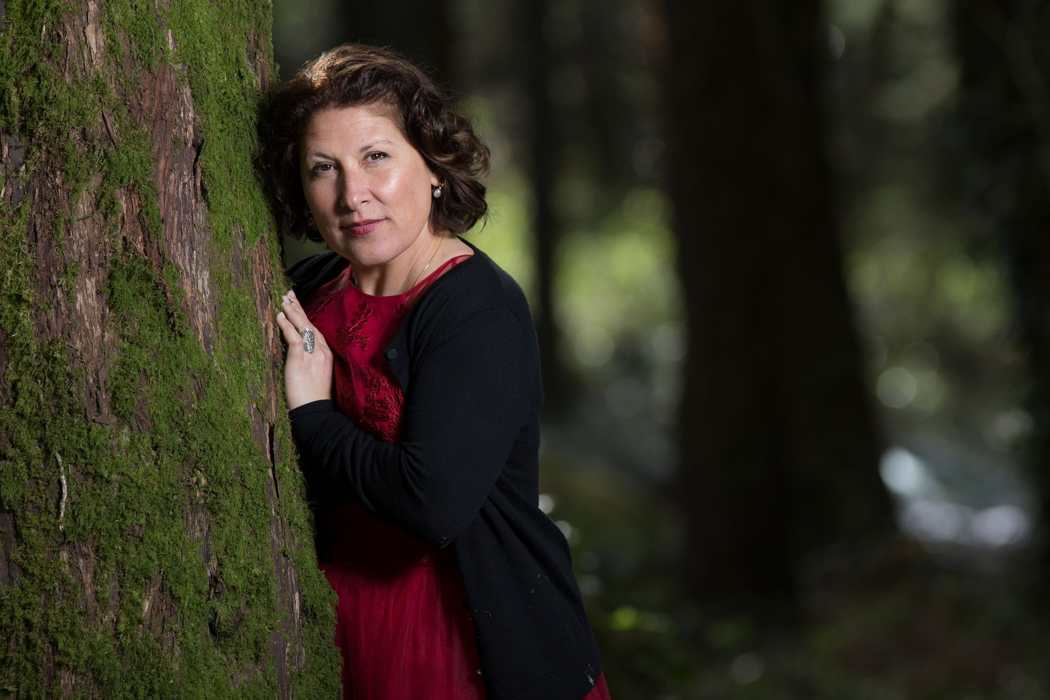 A woman in a red dress leans against a mossy tree in a forest setting, creating a serene and natural vibe.