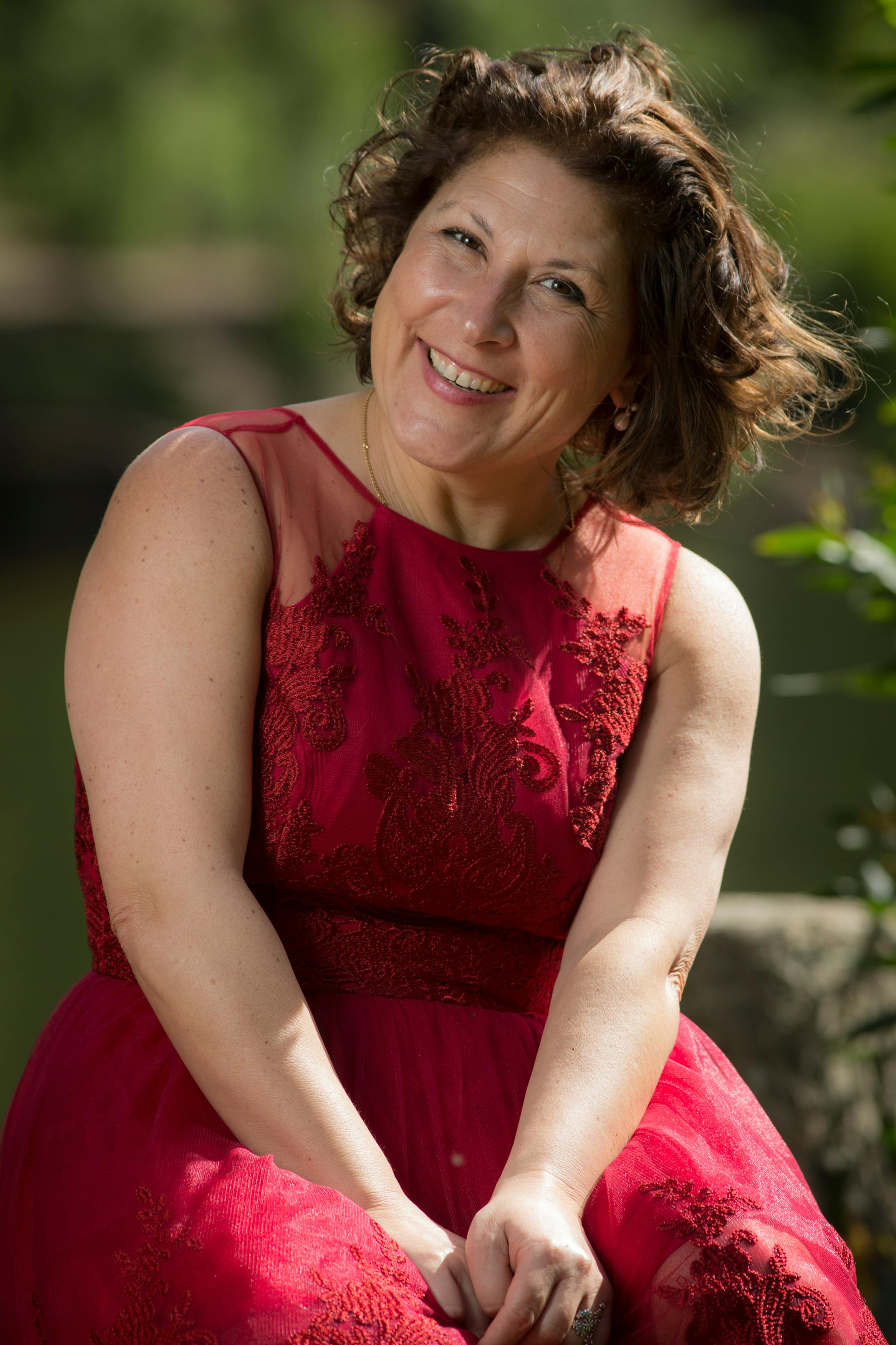 A cheerful woman in a red lace dress smiling under the sunlight.