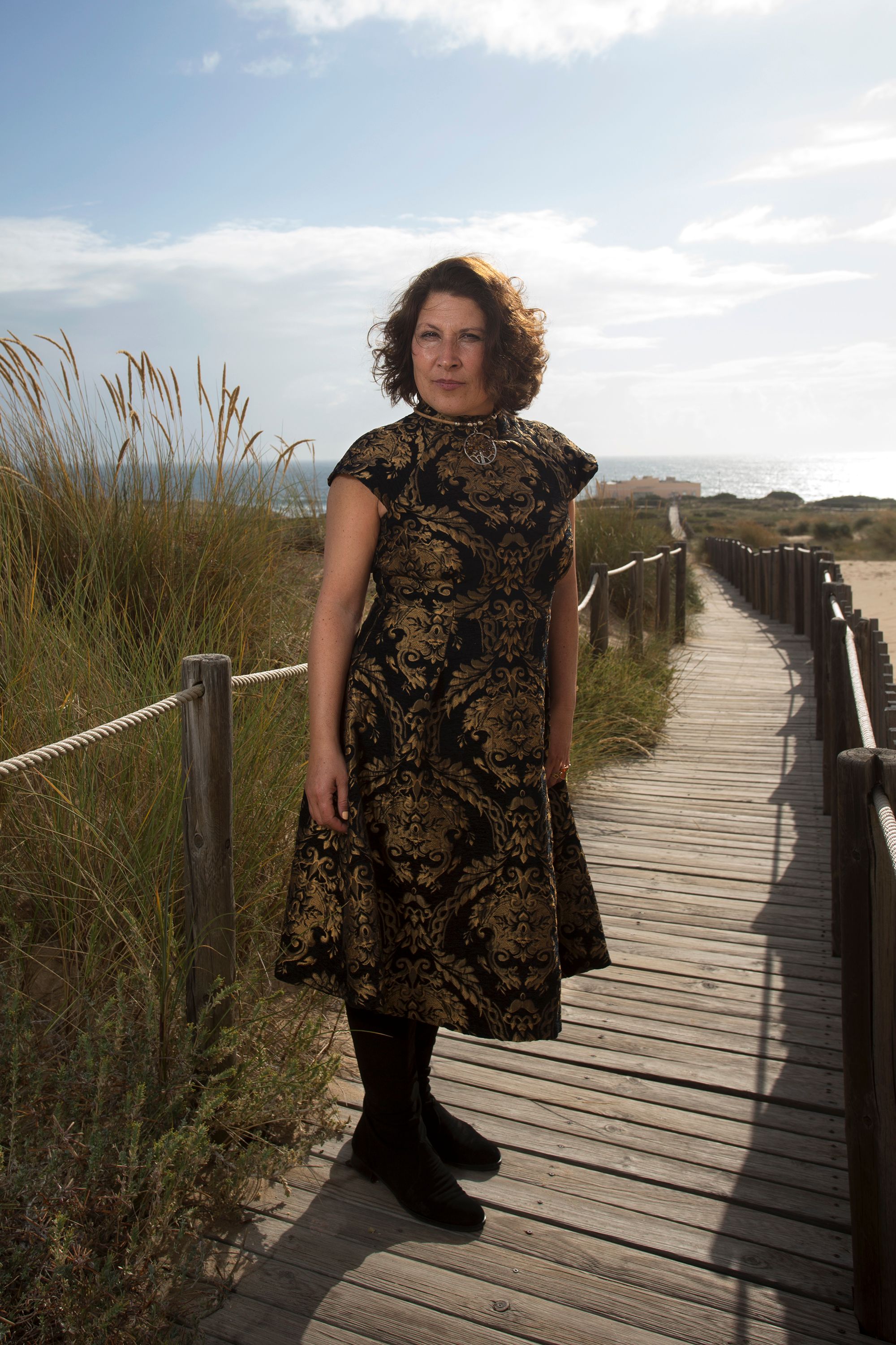 Stylish woman in patterned dress standing on wooden boardwalk by the coast under a sunny sky.
