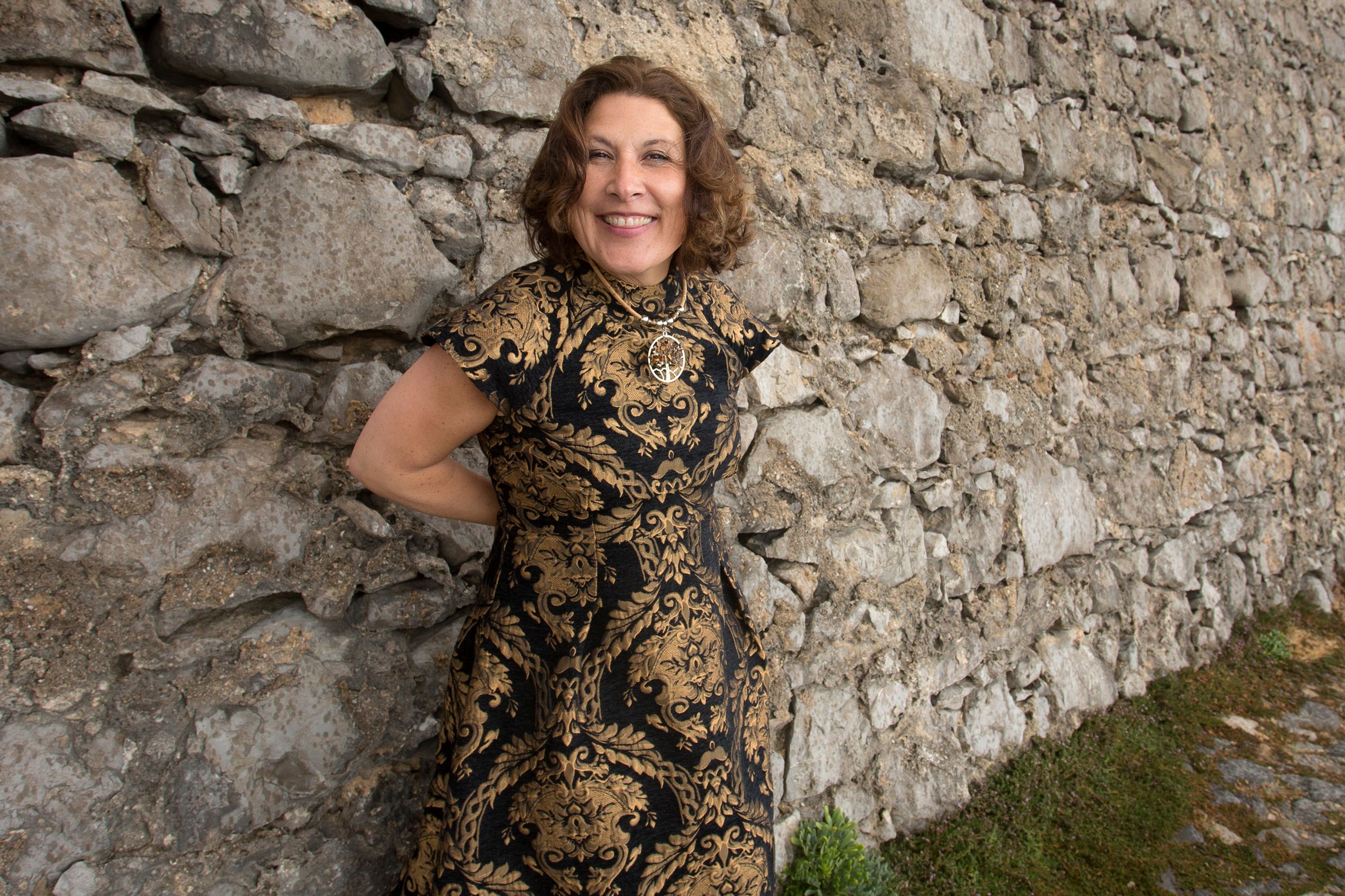 Happy woman in a patterned dress smiles against a rustic stone wall outdoors.