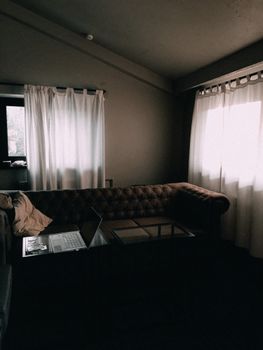 A dimly lit living room features a stylish sofa, laptop, and soft white curtains by the window.