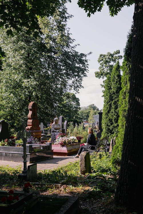 A Person Sitting on a Bench in Front of a Grave with Flowers Near a Statue 