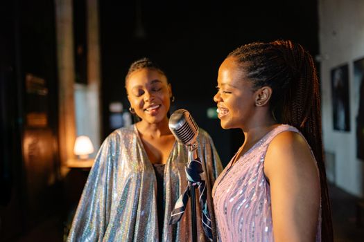 Two women performing indoors, sharing a vintage microphone in joyful harmony.