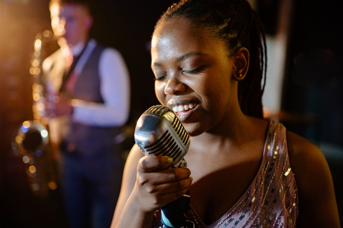 Nyra Okonjo at the microphone, mid-phrase, a saxophonist behind her — photograph by Yan Krukau on Pexels