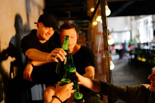 Free Men enjoying a casual gathering, toasting with beer bottles in a lively indoor setting. Stock Photo
