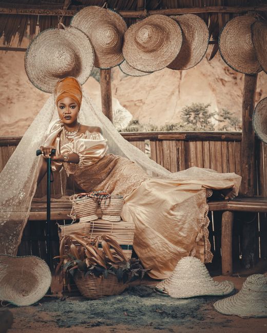 Nigerian woman in traditional clothing and turban seated among woven baskets and hats.