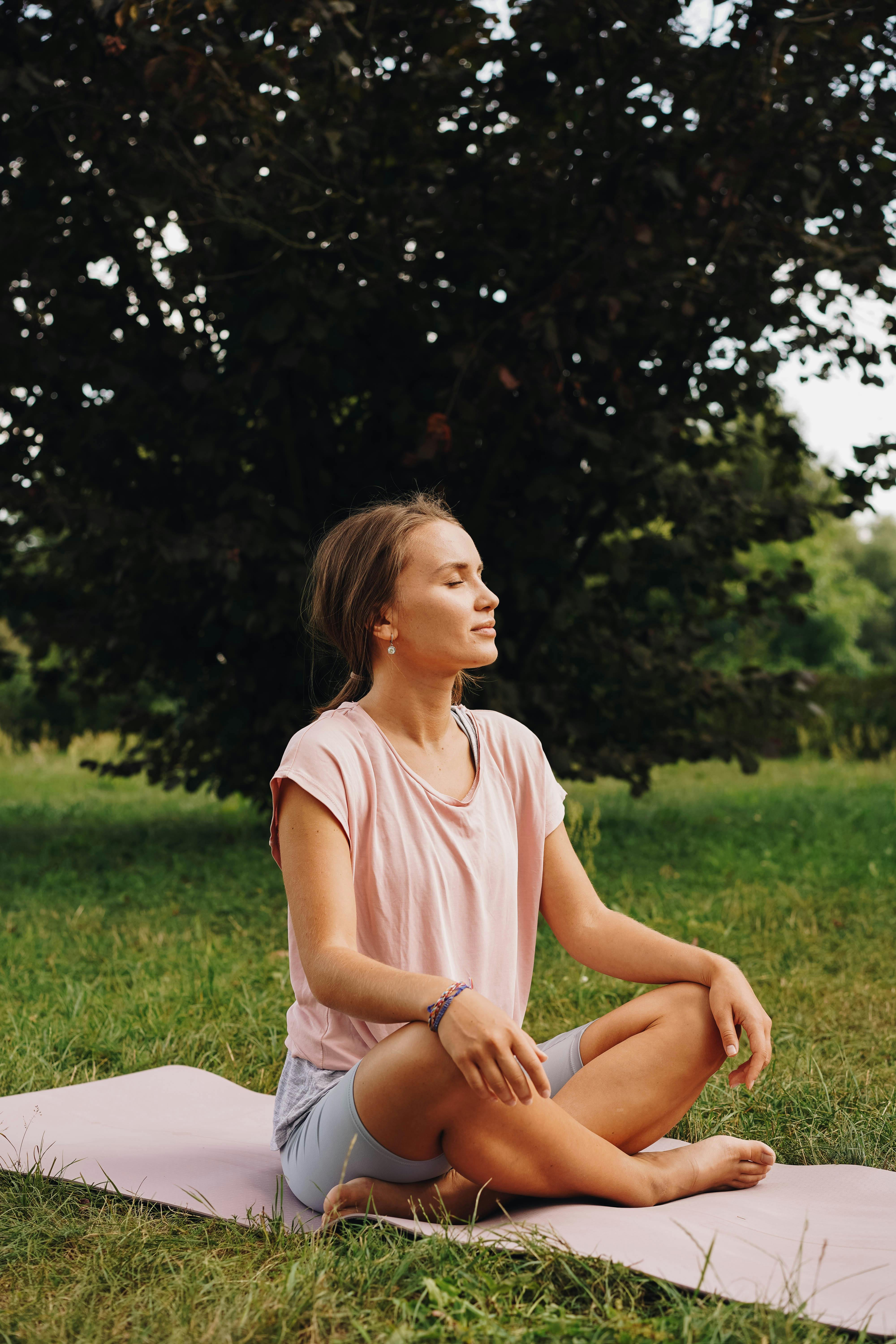 A woman sits peacefully on a yoga mat outdoors, practicing meditation in a serene park setting.