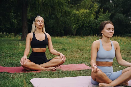 Two women meditating on yoga mats in a tranquil park, promoting mindfulness and relaxation.