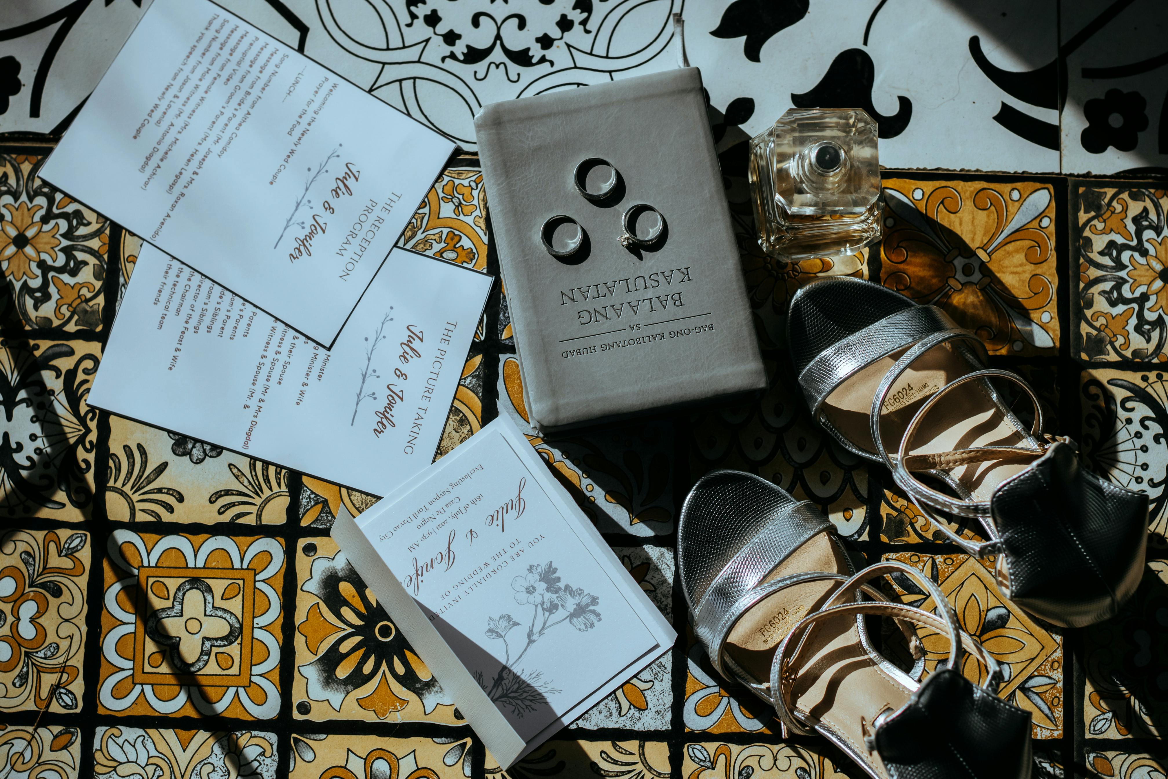 A flat lay of wedding invitations, rings, and shoes on decorative tiles with sunlight.