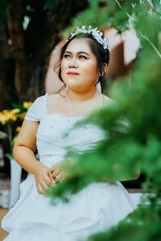 Beautiful bride in a white dress posing elegantly outdoors among greenery.