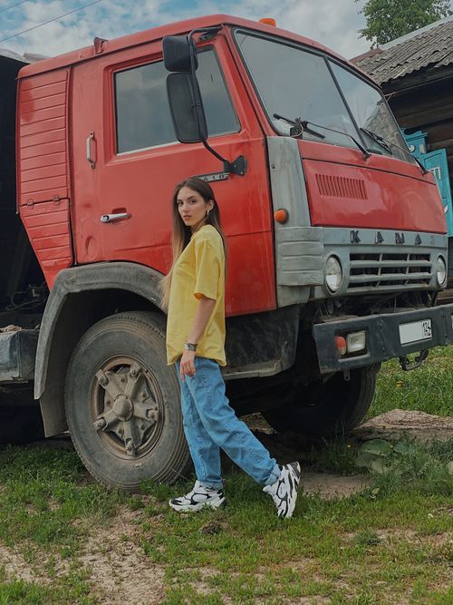Free A young woman in casual wear stands confidently beside a red truck in an outdoor setting. Stock Photo