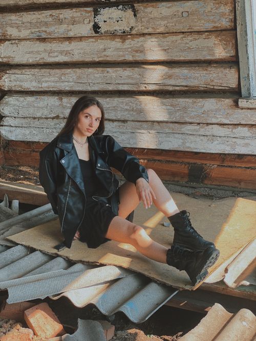 Free Woman in leather jacket sitting on planks against rustic wall in daylight. Stock Photo