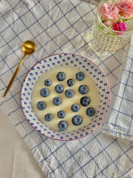 A plate of fresh blueberries in yogurt on a stylish table setting with a gold spoon and floral vase.
