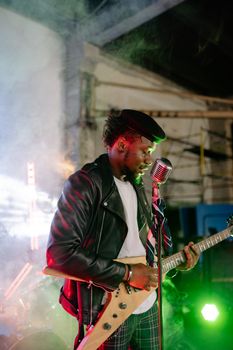 Black musician in leather jacket playing guitar energetically at a night concert.