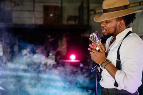 Free A male singer passionately performs indoors with a vintage microphone, wearing a hat and suspenders. Stock Photo