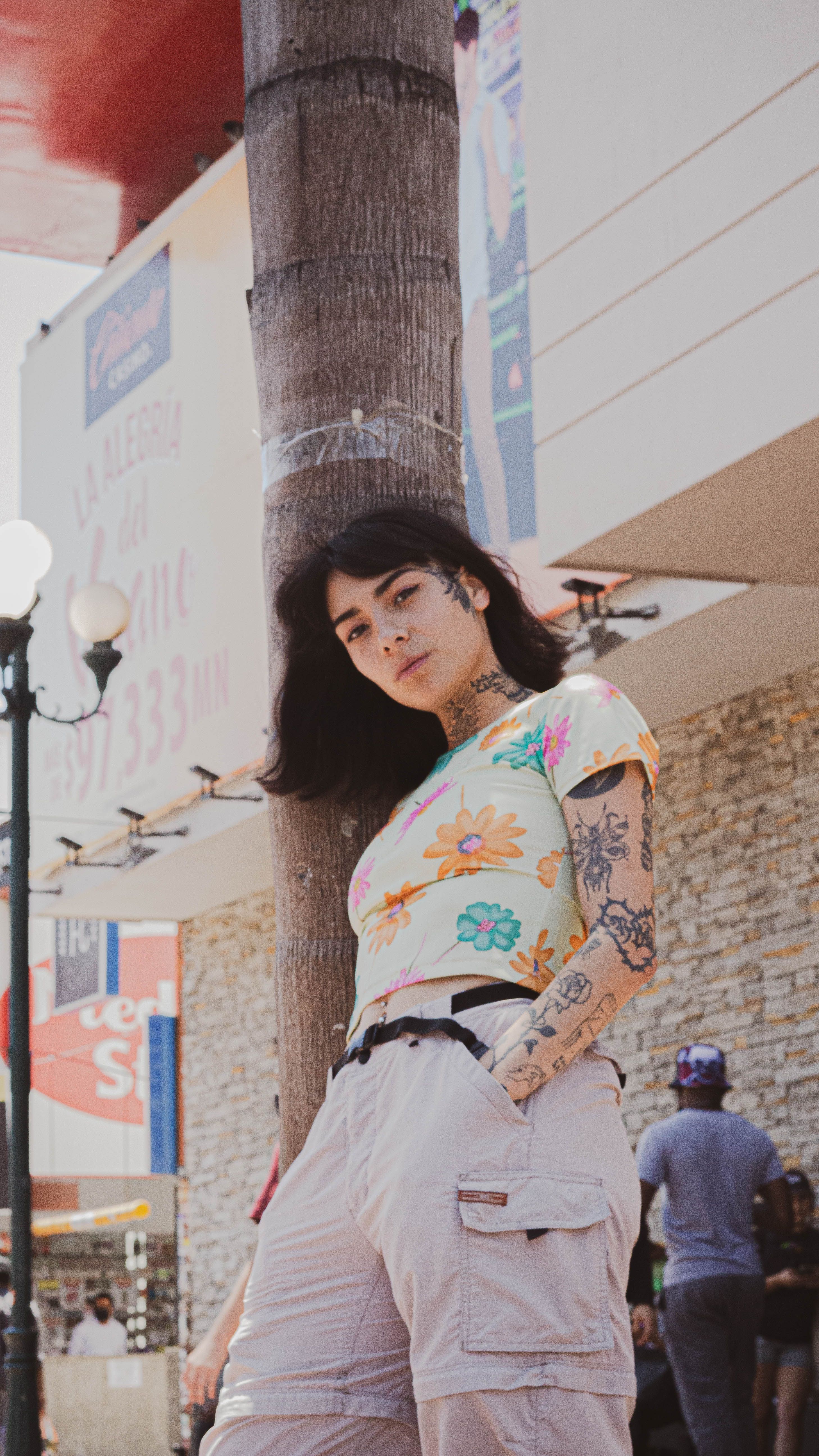 Young tattooed woman in floral top leaning against a palm tree in urban setting on a sunny day.