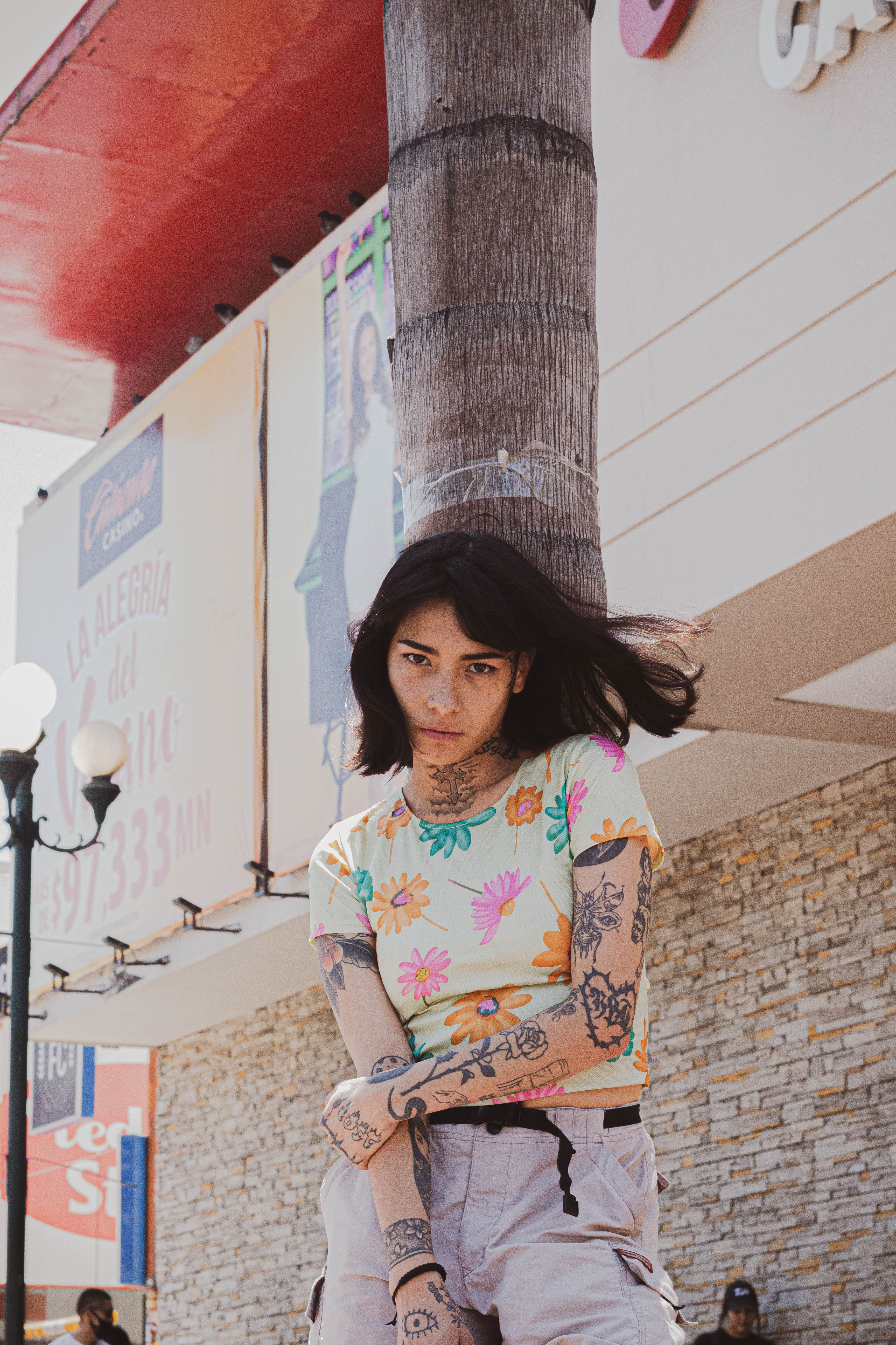 Young tattooed woman in floral top leaning against a palm tree in an urban setting.