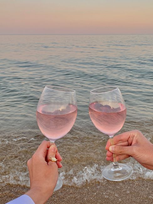 Two people clinking rosé wine glasses on a serene beach at sunset, celebrating a romantic moment.