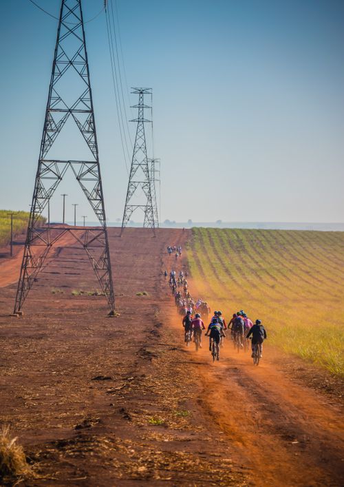 Free Group of cyclists riding on a dirt road beside power lines in a rural landscape during daytime. Stock Photo
