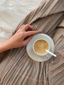 A cozy morning scene with a hand holding a cup of hot coffee on a patterned blanket.