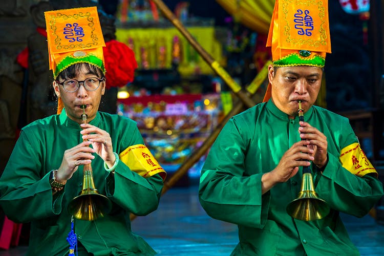 A Pair Of Men In Green Long Sleeve Uniforms With Yellow Hats Playing The Trumpets