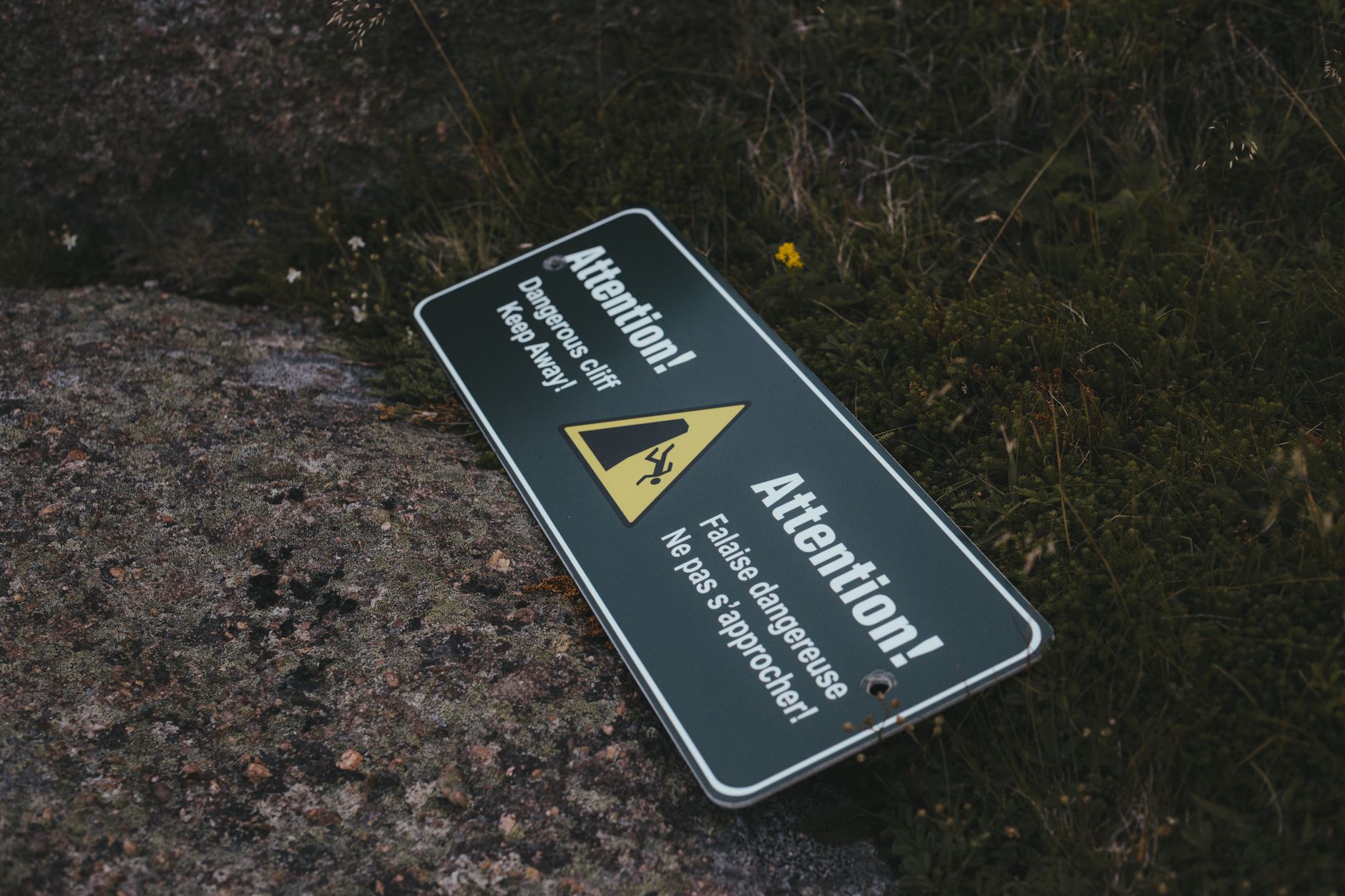 An outdoor warning sign saying 'Attention! Dangerous cliff. Keep away.' lying on rocky ground.