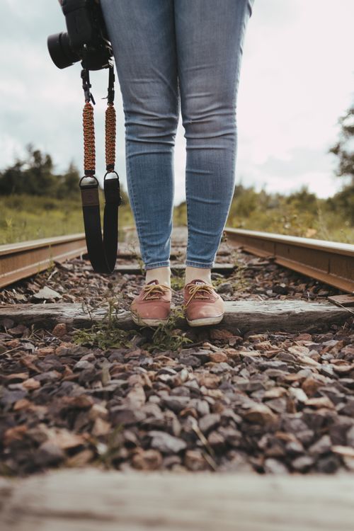 Free Person wearing jeans and sneakers stands on railway tracks holding a camera, signifying a journey or adventure. Stock Photo