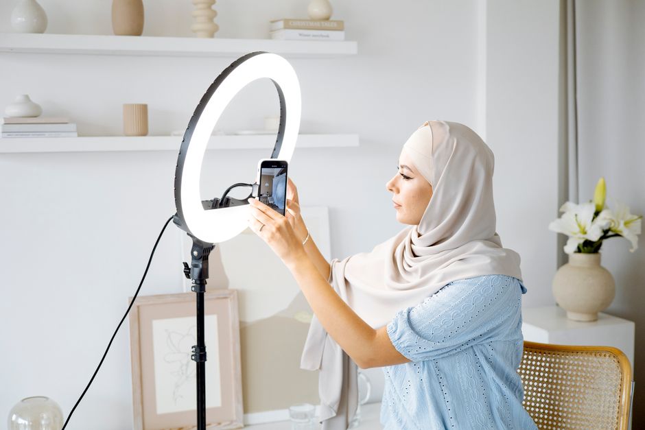Muslim woman in hijab setting up smartphone with ring light in a modern interior.