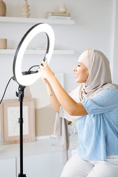 Muslim woman in hijab adjusting a ring light indoors, wearing traditional attire and smiling.