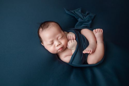 Free Close-up of a peaceful Asian newborn sleeping wrapped in blue cloth against a blue background. Stock Photo