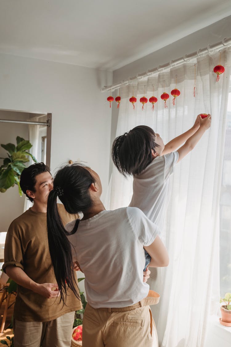 Woman Carrying A Child Reaching Decorations On A White Curtain