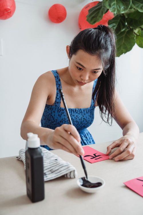 Free Focused woman painting calligraphy on red paper with a brush indoors. Stock Photo