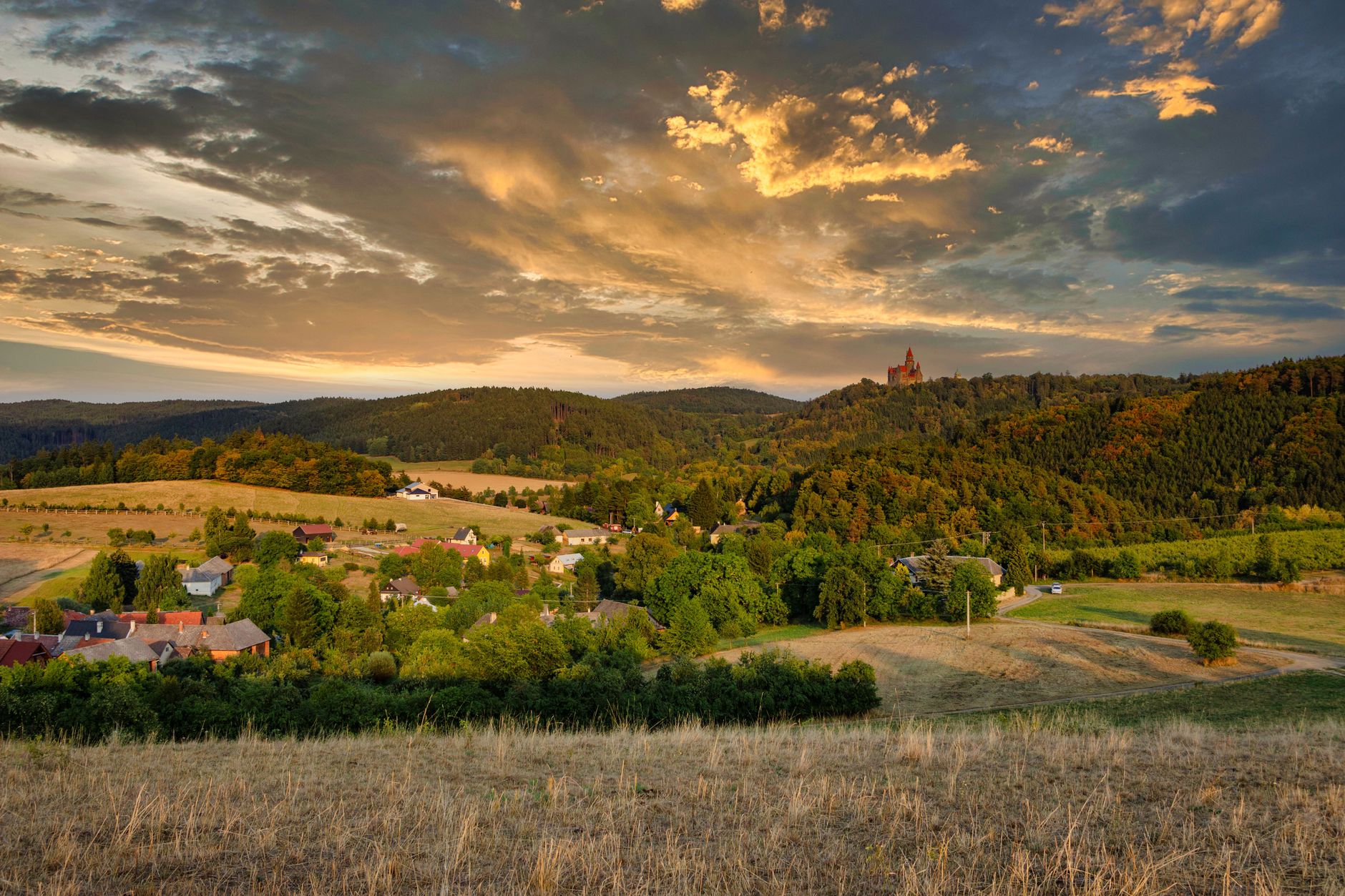 Scenic view of a tranquil Czech village surrounded by lush hills at sunset, under an orange sky.