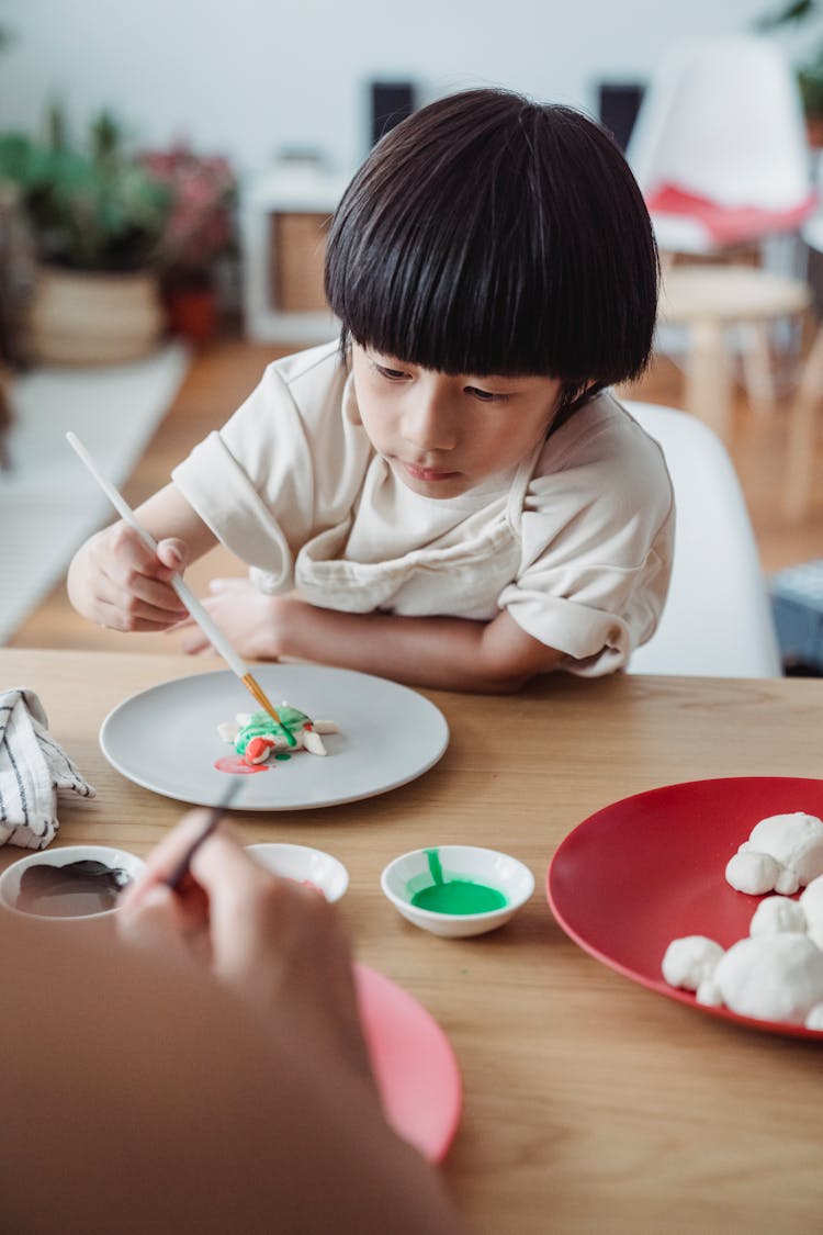 Brunette Boy Sitting At Wooden Table And Colouring Salt Dough 