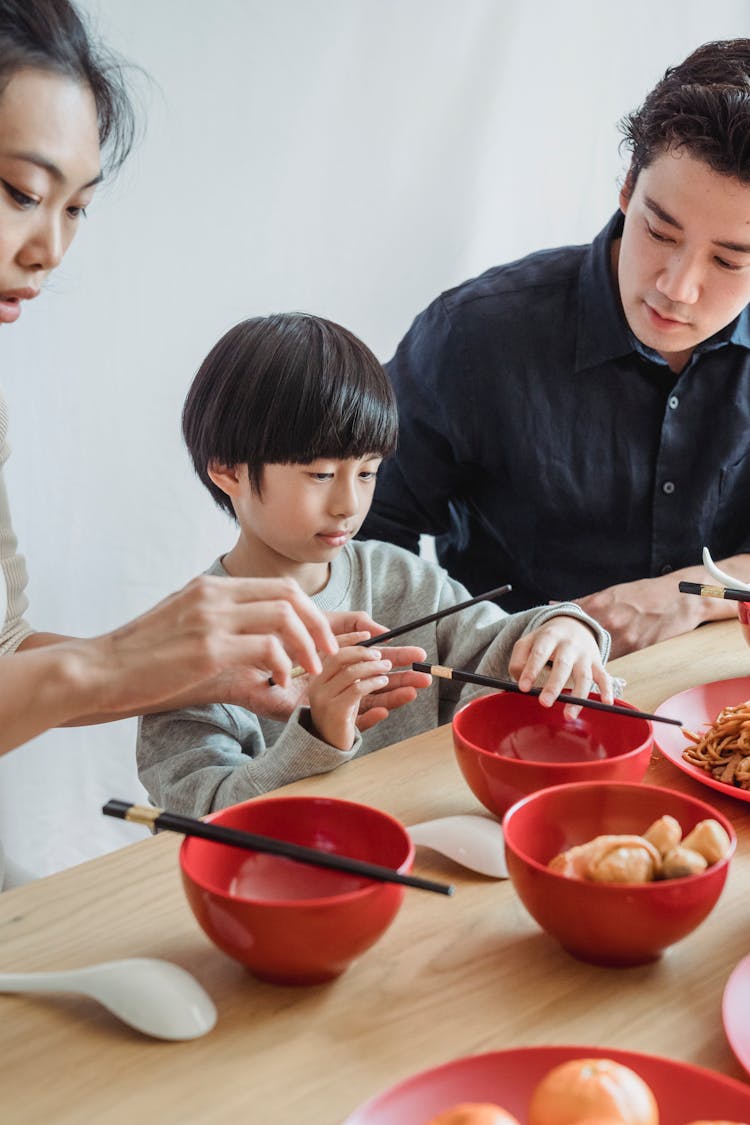 A Child Holding Chopsticks Sitting Between A Man And A Woman 