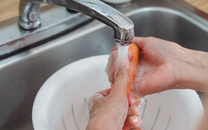 Person washing fresh carrots under a kitchen faucet using a colander, in a home setting.