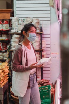 Masked Asian woman shopping in an indoor market, examining products with a thoughtful expression.