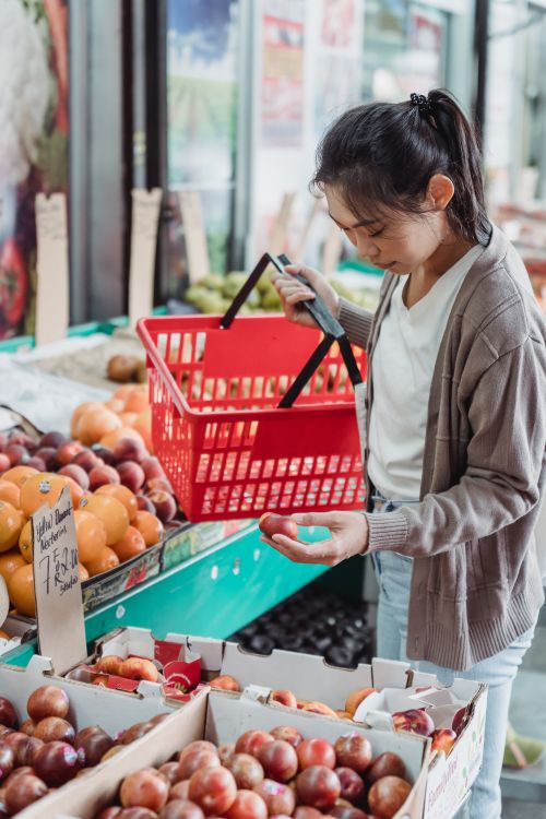 Free A young adult woman shops for fresh fruits at a farmer's market, holding a red basket. Stock Photo