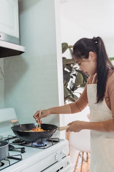 Asian woman cooking with chopsticks in a wok on a stove in a bright kitchen.