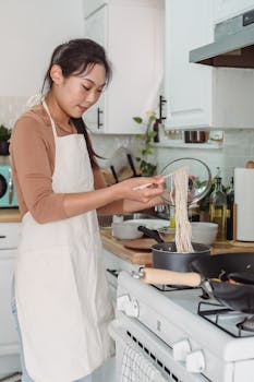 An Asian woman in an apron prepares noodles on a stove in a bright kitchen.