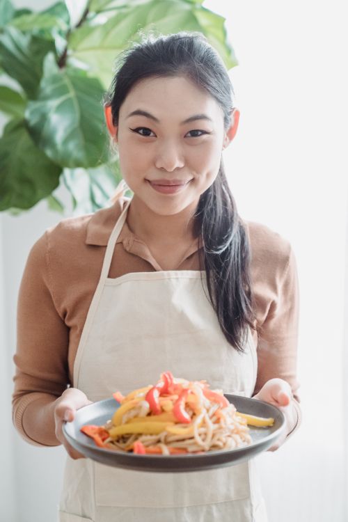 Free Smiling woman presenting a vibrant plate of pasta with vegetables indoors. Stock Photo