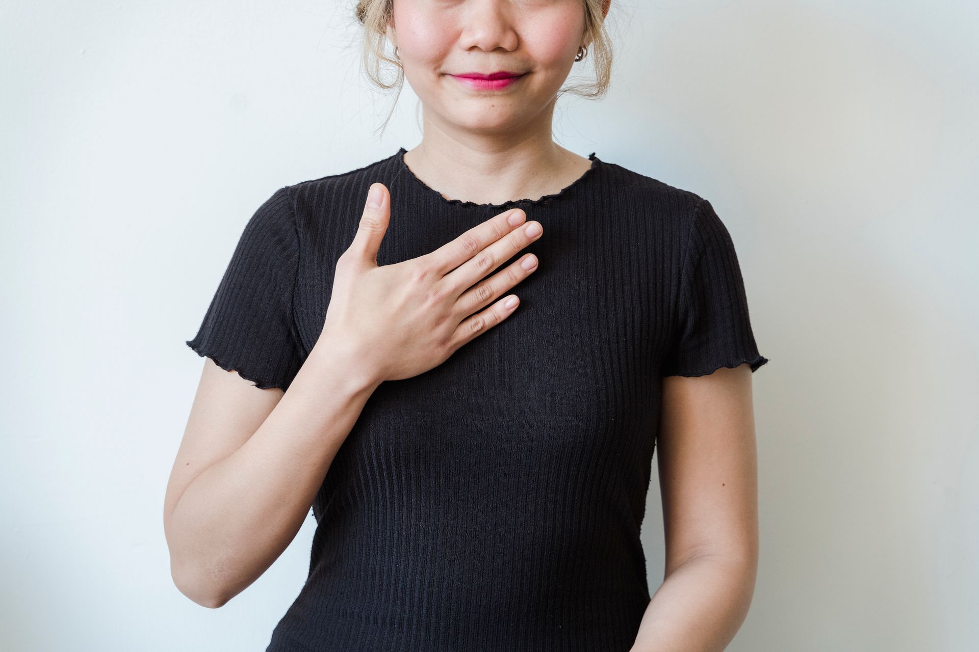 Asian woman in a black shirt expressing gratitude with a hand gesture against a neutral background.