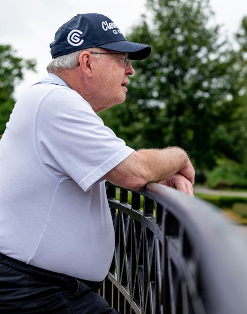 An Elderly Man in Black Cap Wearing Polo Shirt while Leaning by the Metal Railing
